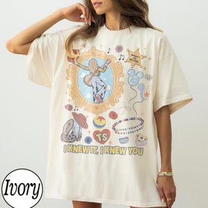 May include: An ivory short-sleeve t-shirt featuring a graphic print of a person playing guitar inside an ornate gold frame, surrounded by illustrations of a star with text "INFINITY AND BEYOND", balloons, a cowboy boot, and "I KNEW IT, I KNEW YOU".