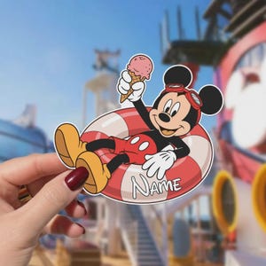 May include: A die-cut vinyl sticker featuring Mickey Mouse wearing red shorts and goggles, lounging in a red and white striped life preserver while holding a pink ice cream cone. The word NAME is printed in white on the life preserver.