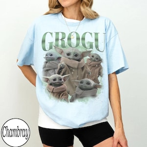 May include: A light blue t-shirt featuring a graphic print of multiple Grogu characters in tan robes. The text GROGU is printed in large green letters above the characters. The design includes subtle white star-like accents and a soft green background.