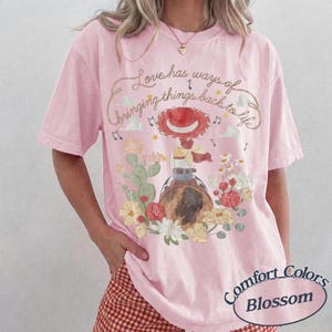 May include: A light pink short-sleeve t-shirt featuring a graphic of a cowgirl on a horse surrounded by cacti and flowers. The text reads, Love has ways of bringing things back to life. Musical notes and stars are scattered around the illustration.