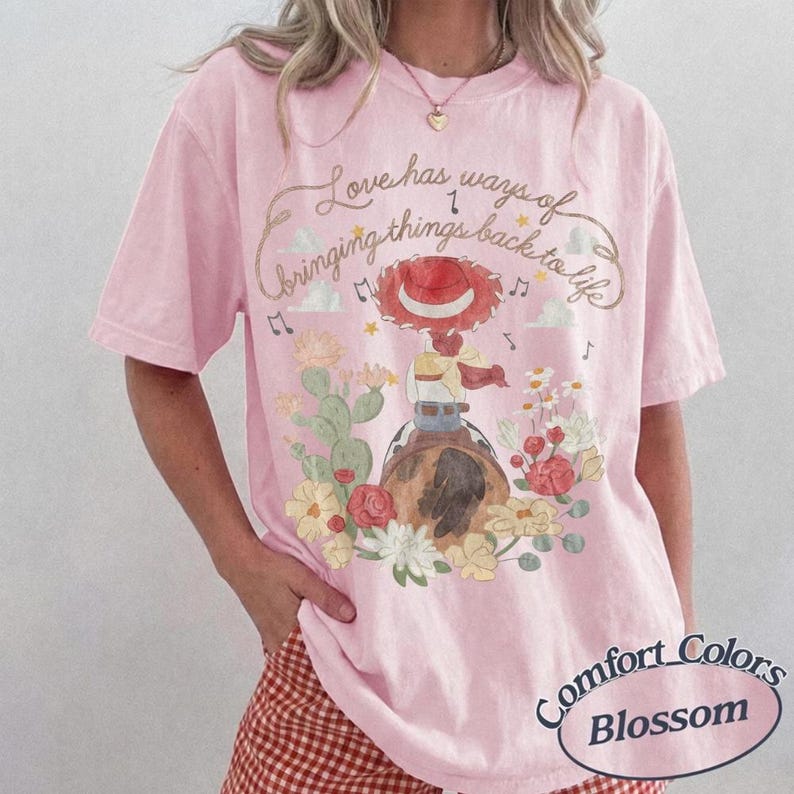 May include: A light pink short-sleeve t-shirt featuring a graphic of a cowgirl on a horse surrounded by cacti and flowers. The text reads, Love has ways of bringing things back to life. Musical notes and stars are scattered around the illustration.
