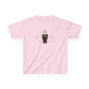 May include: A pale pink t-shirt with a graphic of a Guinness pint, a crown, and arms. The Guinness pint is dark brown with the Guinness logo. The arms are holding the pint. The t-shirt has a crew neck.