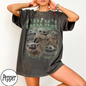 May include: A charcoal gray short-sleeve t-shirt featuring a graphic print of multiple Grogu characters in various poses. The text GROGU is printed in large, bold, light green letters above the characters. The shirt has a relaxed, loose fit.