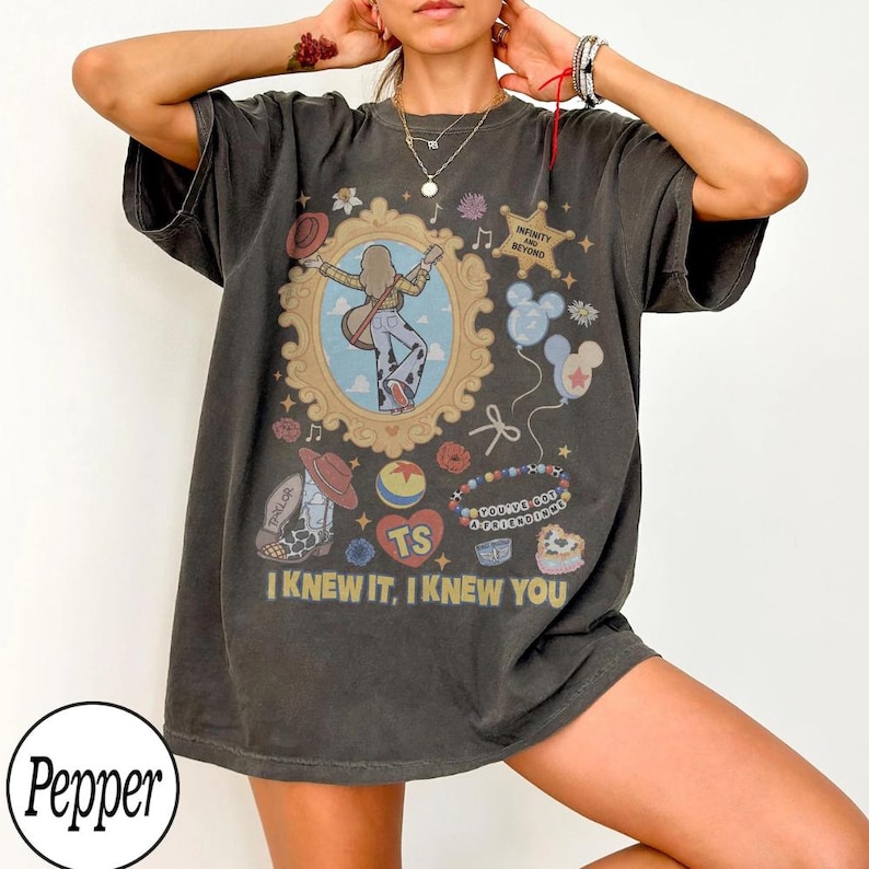 May include: A charcoal gray short-sleeve t-shirt featuring a graphic print with a central ornate gold frame containing a figure with a guitar, surrounded by icons including a star, balloons, a cowboy boot, and text reading I KNEW IT, I KNEW YOU.