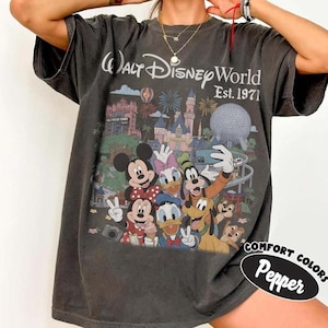 May include: A charcoal grey t-shirt featuring a graphic print of Walt Disney World Est. 1971 with Mickey Mouse, Minnie Mouse, Donald Duck, Daisy Duck, Goofy, Pluto, Chip, and Dale taking a selfie in front of the park landmarks and fireworks.