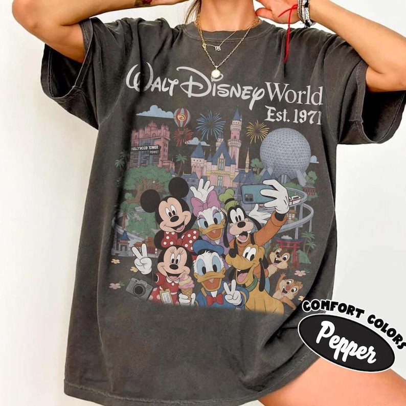 May include: A charcoal grey t-shirt featuring a graphic print of Walt Disney World Est. 1971 with Mickey Mouse, Minnie Mouse, Donald Duck, Daisy Duck, Goofy, Pluto, Chip, and Dale taking a selfie in front of the park landmarks and fireworks.