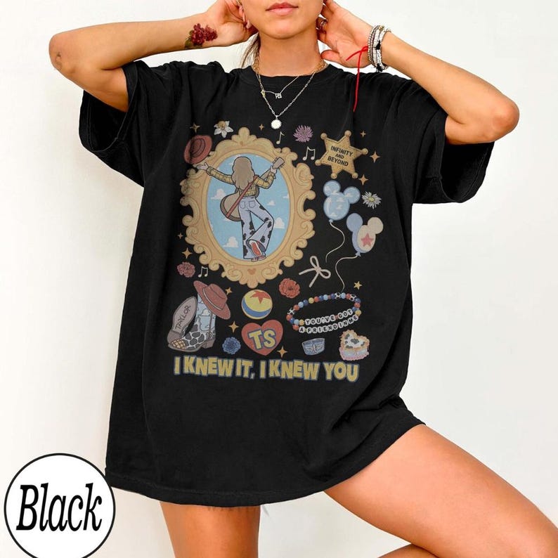 May include: A black short-sleeve t-shirt featuring a graphic print with a central ornate gold frame containing a figure with a guitar. Surrounding elements include a star with text, balloons, a ball, cowboy boots, and the text I KNEW IT, I KNEW YOU.