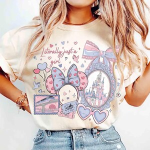 Pode incluir: Camiseta creme com um design extravagante com o texto "Literally just a girl". O gráfico inclui um castelo, óculos de sol em forma de coração, orelhas da Minnie Mouse e outros elementos fofos em rosa e azul.
