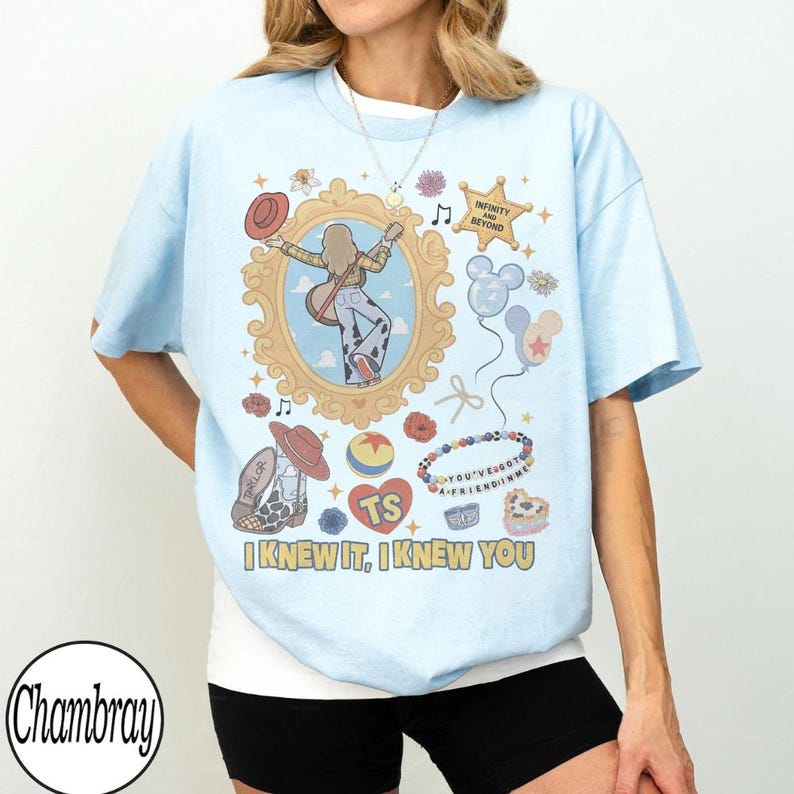May include: A light blue t-shirt featuring a graphic print with a central ornate gold frame containing a figure with a guitar, surrounded by various icons including a star, balloons, a beaded bracelet, and the text "I KNEW IT, I KNEW YOU".