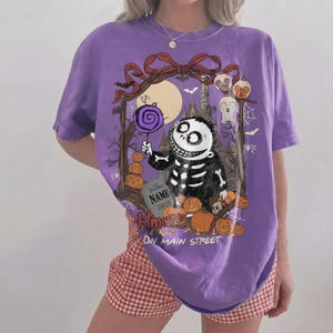 Może przedstawiać: Fioletowy t-shirt z kr&oacute;tkim rękawem z grafiką przedstawiającą szkielet trzymający fioletowego lizaka. Wz&oacute;r zawiera dynie, nagrobek z napisem NAME 2026 oraz tekst Htmare ON MAIN STREET umieszczony w ozdobnej ramce.