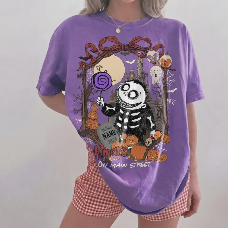 Może przedstawiać: Fioletowy t-shirt z kr&oacute;tkim rękawem z grafiką przedstawiającą szkielet trzymający fioletowego lizaka. Wz&oacute;r zawiera dynie, nagrobek z napisem NAME 2026 oraz tekst Htmare ON MAIN STREET umieszczony w ozdobnej ramce.