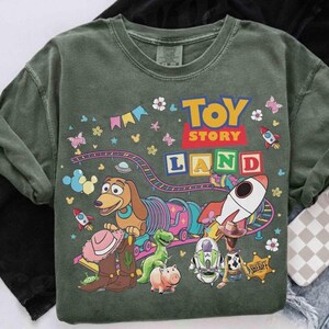 Pode incluir: T-shirt verde de gola redonda com um gr&aacute;fico "Toy Story Land". O design inclui personagens de desenhos animados, foguetes e o texto "Toy Story Land". A camisa tem um visual vintage desbotado.