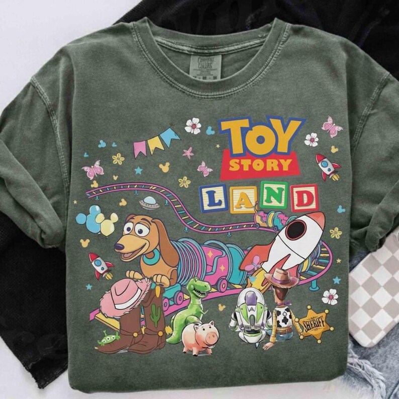 Pode incluir: T-shirt verde de gola redonda com um gr&aacute;fico "Toy Story Land". O design inclui personagens de desenhos animados, foguetes e o texto "Toy Story Land". A camisa tem um visual vintage desbotado.