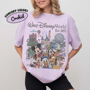 May include: A light purple t-shirt featuring a graphic print of Walt Disney World Est. 1971 with Mickey Mouse, Minnie Mouse, Donald Duck, Daisy Duck, Goofy, Pluto, Chip, and Dale in front of the castle and Epcot ball. A small logo reads Comfort Colors Orchid.