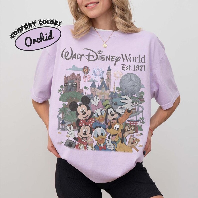 May include: A light purple t-shirt featuring a graphic print of Walt Disney World Est. 1971 with Mickey Mouse, Minnie Mouse, Donald Duck, Daisy Duck, Goofy, Pluto, Chip, and Dale in front of the castle and Epcot ball. A small logo reads Comfort Colors Orchid.