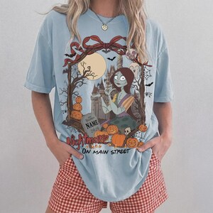 Może przedstawiać: Jasnoniebieski t-shirt z nadrukiem graficznym przedstawiającym postać kobiecą z długimi brązowymi włosami, zamek, pełnię księżyca, nietoperze, pajęczyny i dynie. Tekst brzmi "NIGHTMARE ON MAIN STREET" i zawiera nagrobek z napisem "NAME 2026".