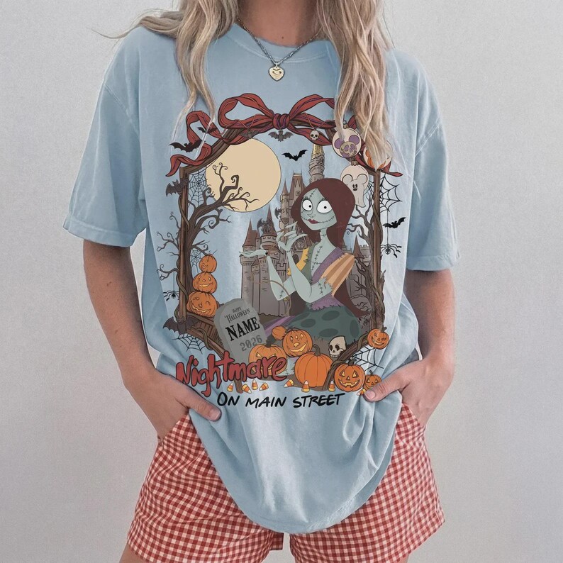 Może przedstawiać: Jasnoniebieski t-shirt z nadrukiem graficznym przedstawiającym postać kobiecą z długimi brązowymi włosami, zamek, pełnię księżyca, nietoperze, pajęczyny i dynie. Tekst brzmi "NIGHTMARE ON MAIN STREET" i zawiera nagrobek z napisem "NAME 2026".