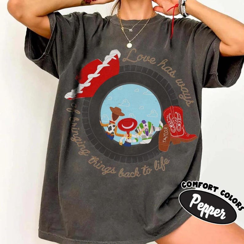 May include: A charcoal gray short-sleeve t-shirt featuring a circular graphic with Toy Story characters Woody, Jessie, and Buzz Lightyear. The design includes a red sneaker, a red cowboy boot, and the text Love has ways of bringing things back to life.