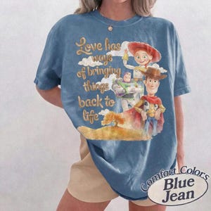 May include: A blue short-sleeve t-shirt featuring a graphic print of characters from Toy Story, including Woody, Buzz Lightyear, and Jessie. The text reads, Love has ways of bringing things back to life, set against a background of clouds and a hill.