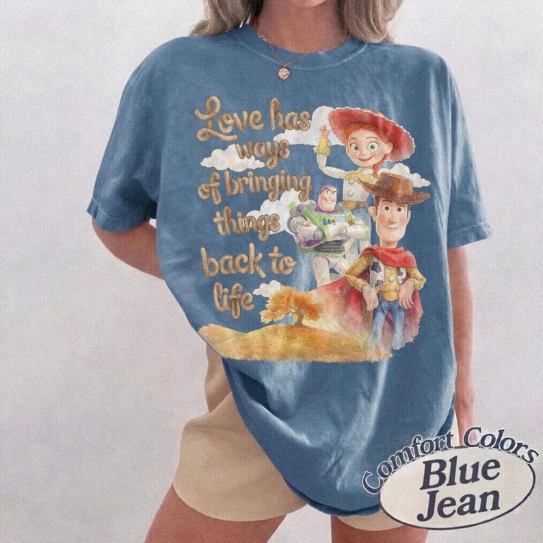 May include: A blue short-sleeve t-shirt featuring a graphic print of characters from Toy Story, including Woody, Buzz Lightyear, and Jessie. The text reads, Love has ways of bringing things back to life, set against a background of clouds and a hill.