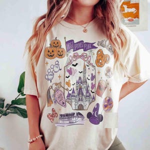 Pode incluir: Uma t-shirt de cor creme com uma impressão gráfica com o texto "MOST SPOOKIEST PLACE". O design inclui um castelo, abóboras, um monotrilho, um chapéu de feiticeiro, orelhas de rato e vários ícones festivos em tons de roxo, laranja e preto.