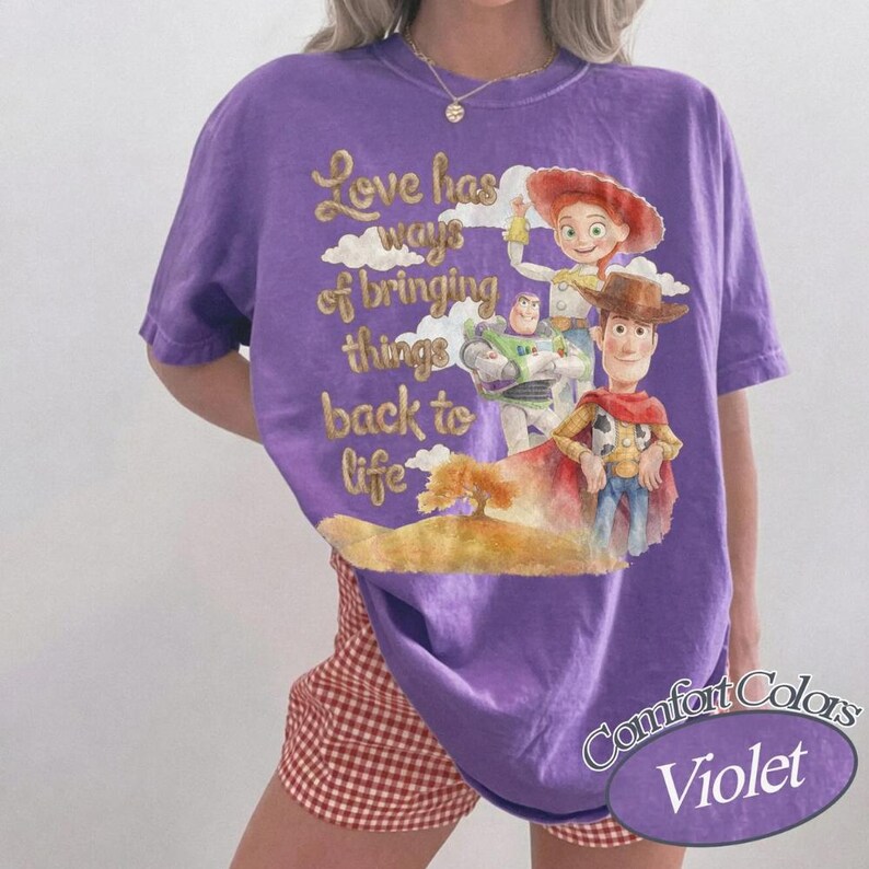 May include: A violet short-sleeve t-shirt featuring a graphic print of Woody, Buzz Lightyear, and Jessie from Toy Story. The text reads, Love has ways of bringing things back to life. The design includes clouds and a landscape in watercolor style.