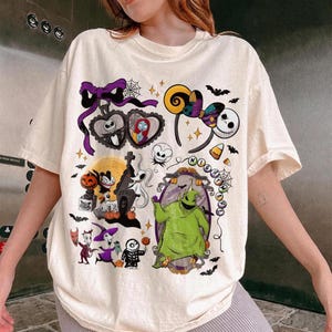 May include: A cream-coloured t-shirt with a colourful collage of Nightmare Before Christmas characters. The design includes Jack Skellington, Sally, Oogie Boogie, and Zero, along with bats, spiderwebs, and other Halloween-themed elements.