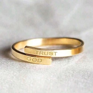May include: A gold-toned ring with the words "TRUST GOD" engraved on it. The ring is made of two bands that are intertwined.
