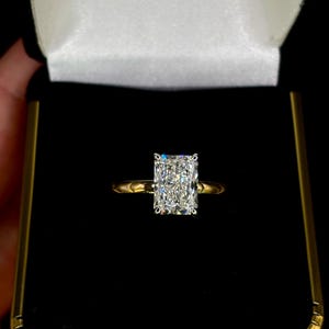 May include: A radiant-cut diamond ring with a gold band, presented in a black and gold jewelry box. The diamond is rectangular with a brilliant sparkle. The ring is set against a black background.