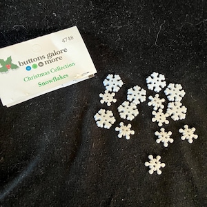 May include: A package of white snowflake buttons labeled "buttons galore & more Christmas Collection Snowflakes" with the number 4748. The package is on a black surface with a scattering of white snowflake buttons.