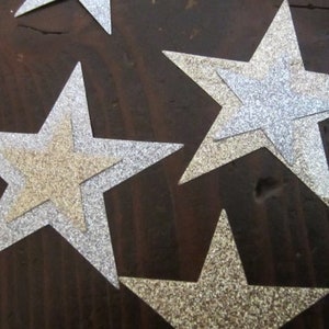 Op de afbeelding: Vier zilveren glittersterren met een gouden glitterster er bovenop gelegd. De sterren zijn in een cluster op een donkerbruin houten oppervlak gerangschikt.