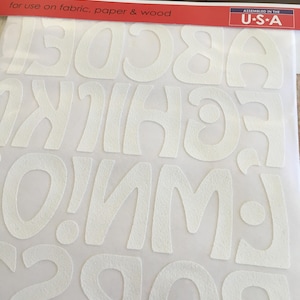 May include: White iron-on letters for crafts. The letters are in a playful, rounded font. The letters are flocked, giving them a soft, fuzzy texture. The package says "iron-on art flocked transfer" and "for use on fabric, paper & wood".