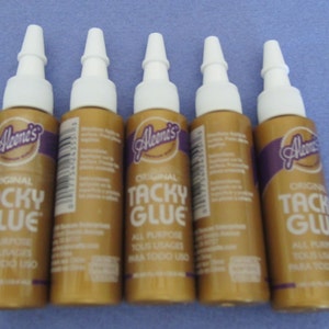 May include: Five bottles of Aleene's Original Tacky Glue. The bottles are gold with a purple label and white caps. The label says "Original Tacky Glue" and "All Purpose, Acid Free, Non Toxic Use."