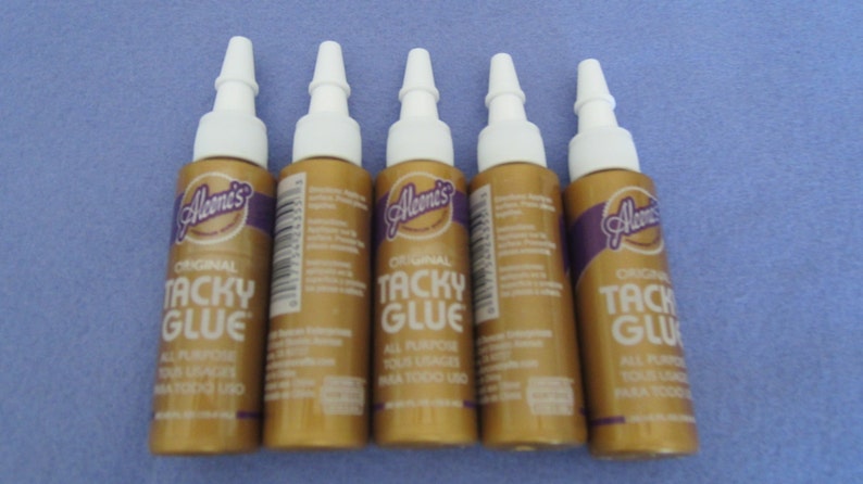 May include: Five bottles of Aleene's Original Tacky Glue. The bottles are gold with a purple label and white caps. The label says "Original Tacky Glue" and "All Purpose, Acid Free, Non Toxic Use."