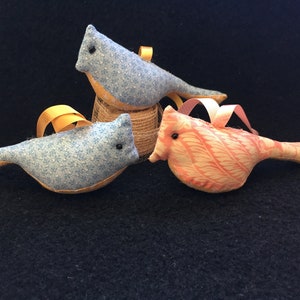 May include: Three handmade fabric bird ornaments. One is blue with white polka dots, one is light blue with white dots, and one is pink with a white leaf pattern. Each bird has a yellow ribbon for hanging.