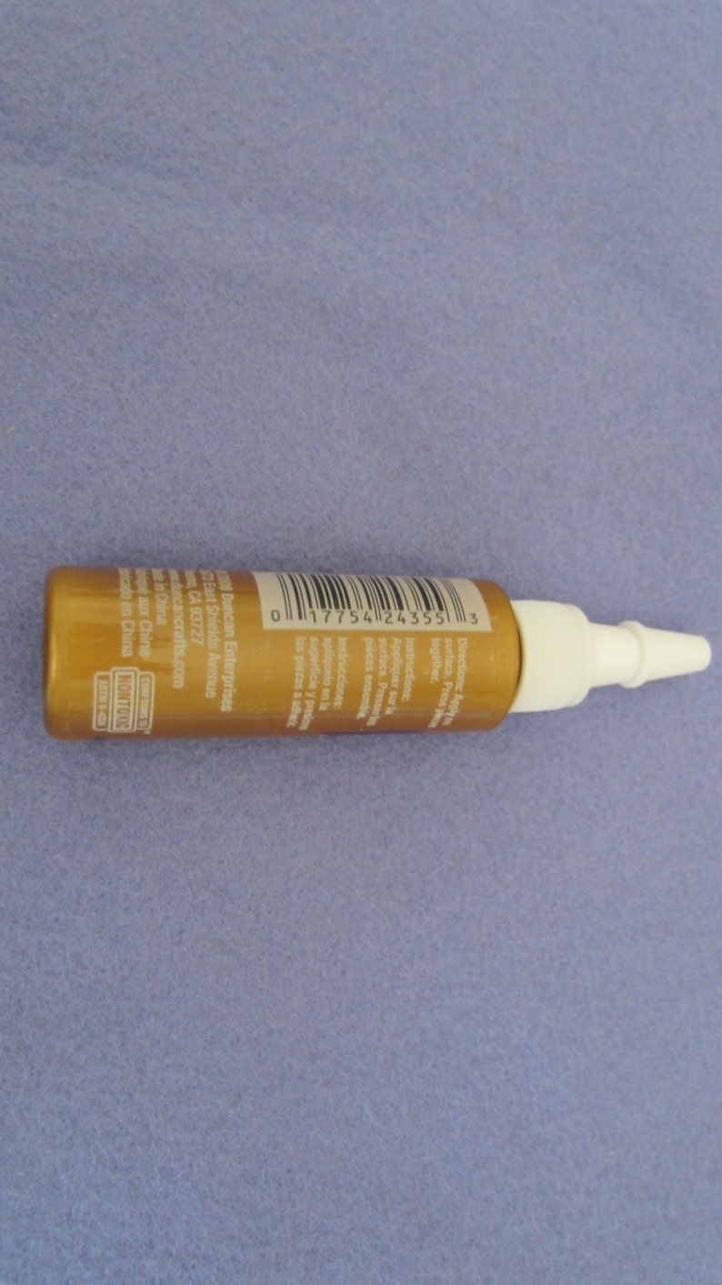 May include: A gold bottle with a white nozzle and a barcode label. The label includes the text "Duncan Enterprises", "East Shields Avenue", "CA 93727", "duncancrafts.com", "Made in China", and "NONTOXIC".