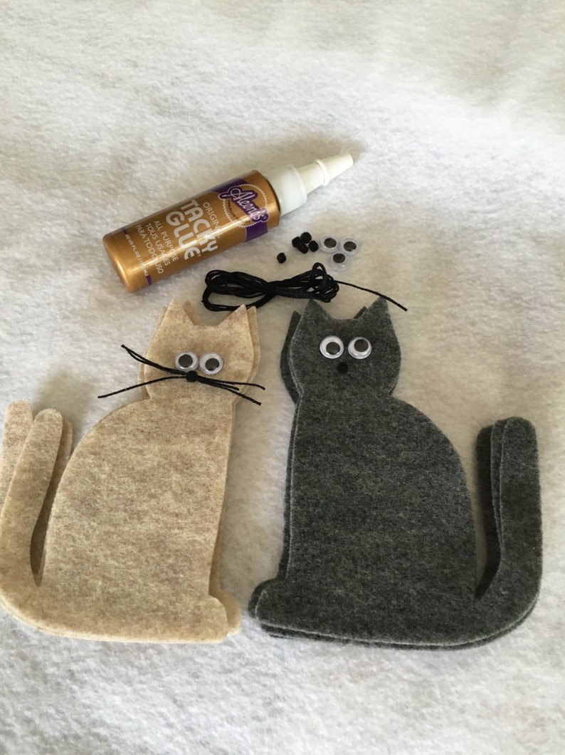 May include: Two felt cat shapes, one beige and one gray, with googly eyes, black yarn whiskers, and black thread for a tail. A bottle of tacky glue, black beads, and extra googly eyes are also included.