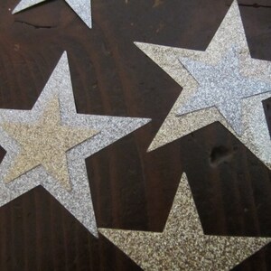 Op de afbeelding: Vier zilveren glittersterren met een gouden glitterster die op een van de zilveren sterren is gelegd. De sterren liggen op een donkerbruin houten oppervlak.