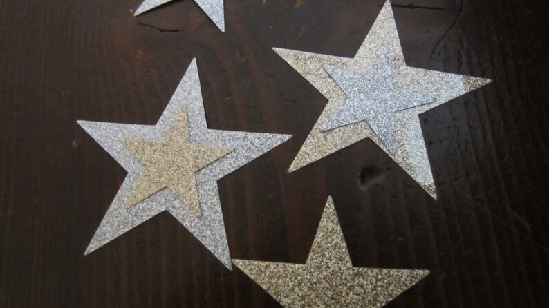 Op de afbeelding: Vier zilveren glittersterren met een gouden glitterster die op een van de zilveren sterren is gelegd. De sterren liggen op een donkerbruin houten oppervlak.