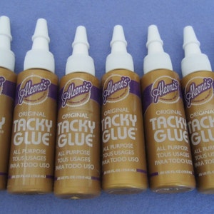 May include: Eight bottles of Aleene's Original Tacky Glue. The bottles are gold with a purple label and white caps. The label says "Aleene's Original Tacky Glue" and "All Purpose Tous Usages Para Todo Uso".