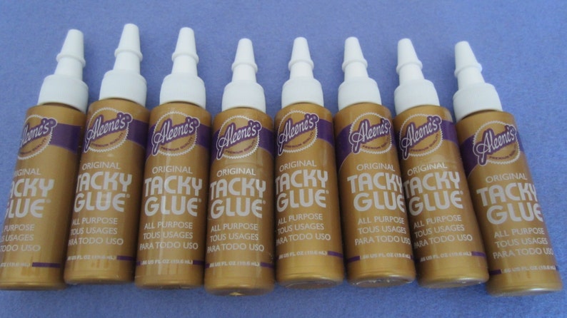 May include: Eight bottles of Aleene's Original Tacky Glue. The bottles are gold with a purple label and white caps. The label says "Aleene's Original Tacky Glue" and "All Purpose Tous Usages Para Todo Uso".