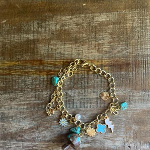 May include: Gold-toned charm bracelet with various pendants, including turquoise stones, a mushroom, a daisy, a four-leaf clover, a lightning bolt, and a sunburst. The bracelet is displayed on a wooden surface.