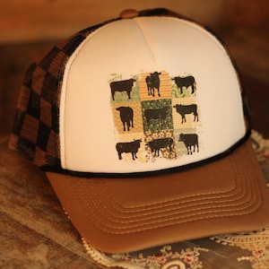 May include: A trucker hat with a brown peak and a white front panel featuring a black and white cow pattern. The hat has a brown and black chequered mesh back and a black rope detail.