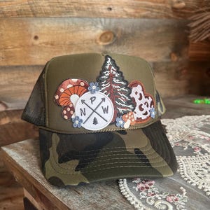 May include: A green trucker hat with a camouflage brim and a mesh back. The front features embroidered patches of mushrooms, a compass rose, a tree, and a cowhide pattern. The hat has a curved brim and a snapback closure.