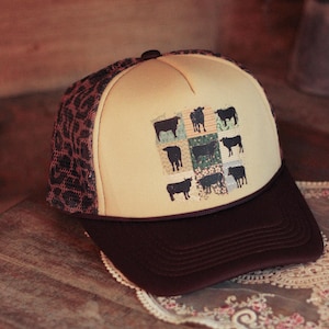 May include: A beige and brown trucker hat with a dark brown peak. The hat features a patch with a collage of black cow illustrations. The mesh backing is a leopard print.