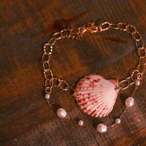 May include: A delicate bracelet featuring a central pink and white scallop shell charm, accented with small white pearls on a thin gold-toned chain. The design includes a larger oval link chain section and a finer chain strand adorned with pearls.