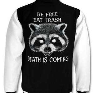 May include: Black and white varsity jacket with white sleeves. The back of the jacket features a raccoon face with the text "Be Free Eat Trash Death is Coming".