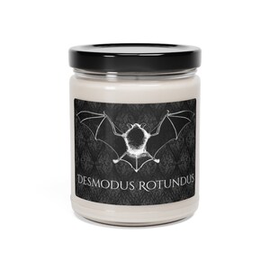 May include: A white candle in a glass jar with a black label. The label features a black and white illustration of a bat with the text "Desmodus Rotundus".