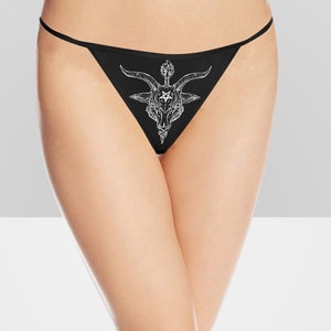 Op de afbeelding: Zwarte kanten string met een witte grafische print van een geitenkop met hoorns en een pentagram.