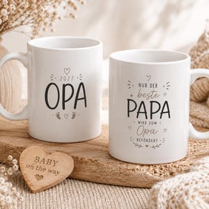 Puede incluir: Dos tazas de cerámica blanca con texto negro y diseños gráficos. Una taza muestra el texto 2027 OPA con iconos de corazones y huellas. La segunda taza presenta el texto NUR DER beste PAPA WIRD ZUM Opa BEFÖRDERT con detalles de corazones y hojas.